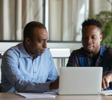 Two African American employees working on project together, using laptop pointing at screen, discussing strategy and sharing ideas at meeting, colleagues involved in teamwork, cooperation concept